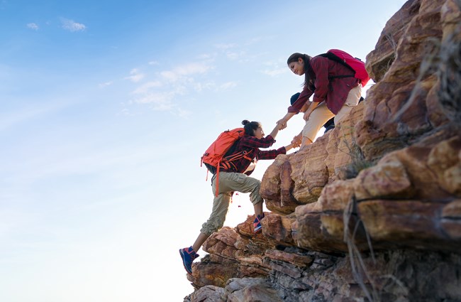2 people rock climbing with one holding out their hand to help the other up.
