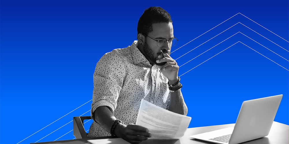 man with glasses and beard holding a piece of paper and looking at his laptop